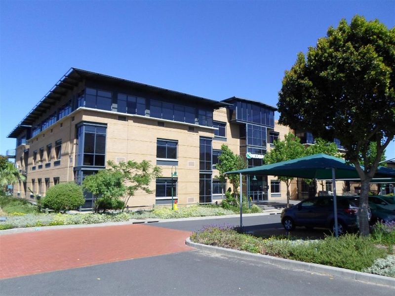 A professional commercial office building featuring a brick facade, extensive glazing, and a paved parking area with shade cloth carports.