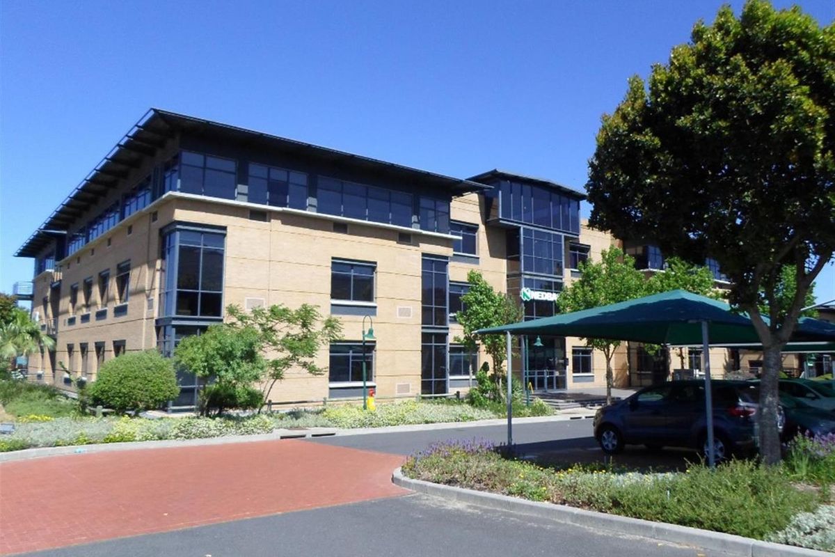 A professional commercial office building featuring a brick facade, extensive glazing, and a paved parking area with shade cloth carports.