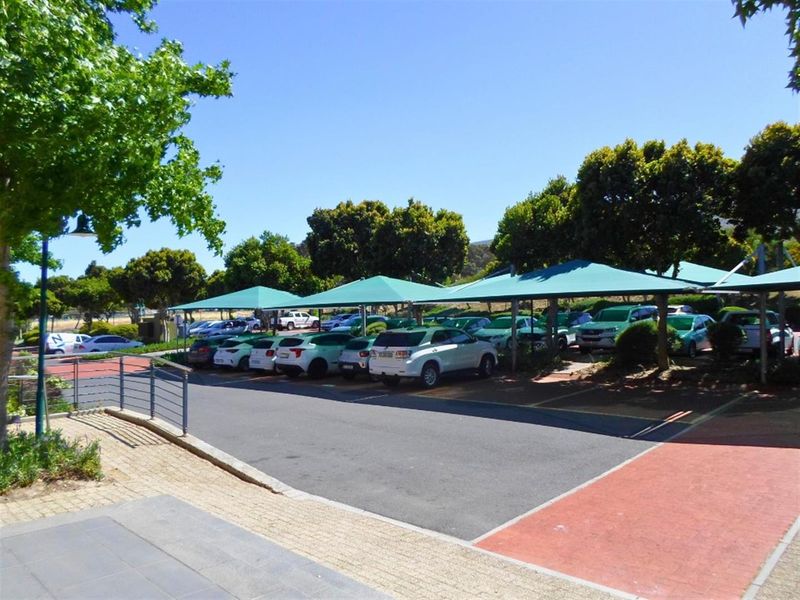 A well-maintained commercial parking area featuring multiple shade cloth carports, asphalt surfacing, and paved pedestrian walkways surrounded by mature trees.