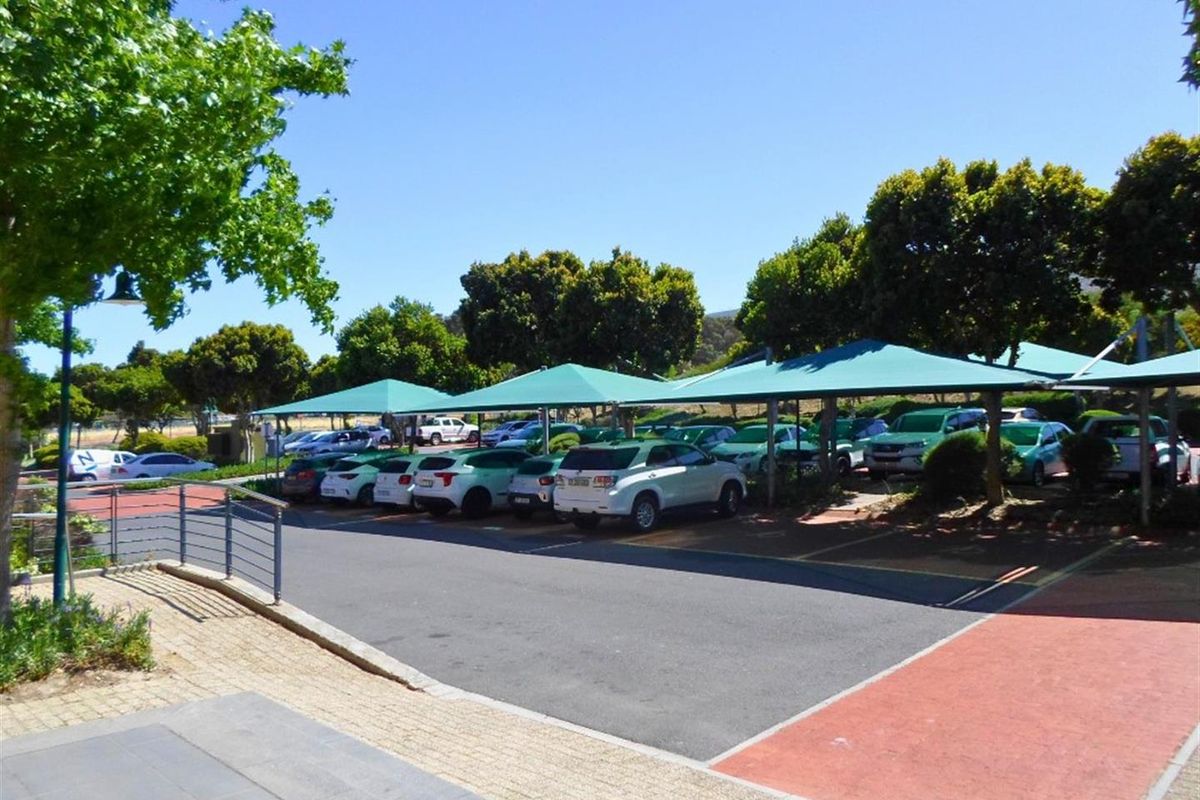 A well-maintained commercial parking area featuring multiple shade cloth carports, asphalt surfacing, and paved pedestrian walkways surrounded by mature trees.