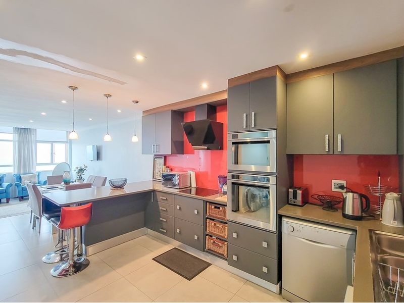 Modern open-plan kitchen featuring dark grey cabinetry, a vibrant red splashback, built-in appliances, and a breakfast bar flowing into the living area.