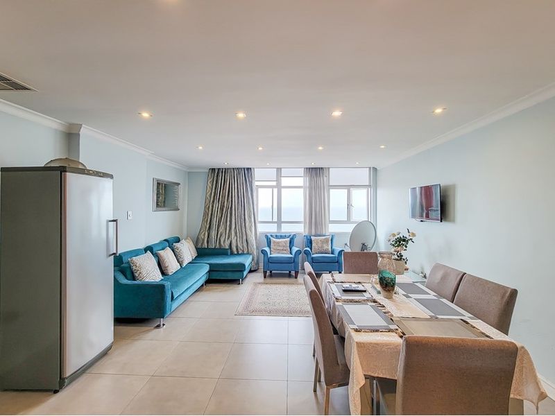 Spacious open-plan living and dining area featuring neutral tiled flooring, recessed downlighting, and large windows providing natural light and ocean views.