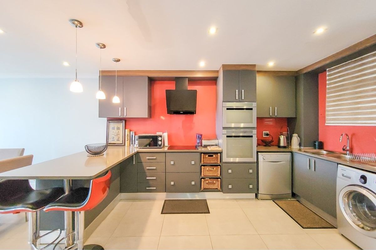 Modern kitchen featuring dark grey cabinetry, a vibrant red splashback, built-in oven, integrated appliances, and a functional breakfast bar with pendant lighting.