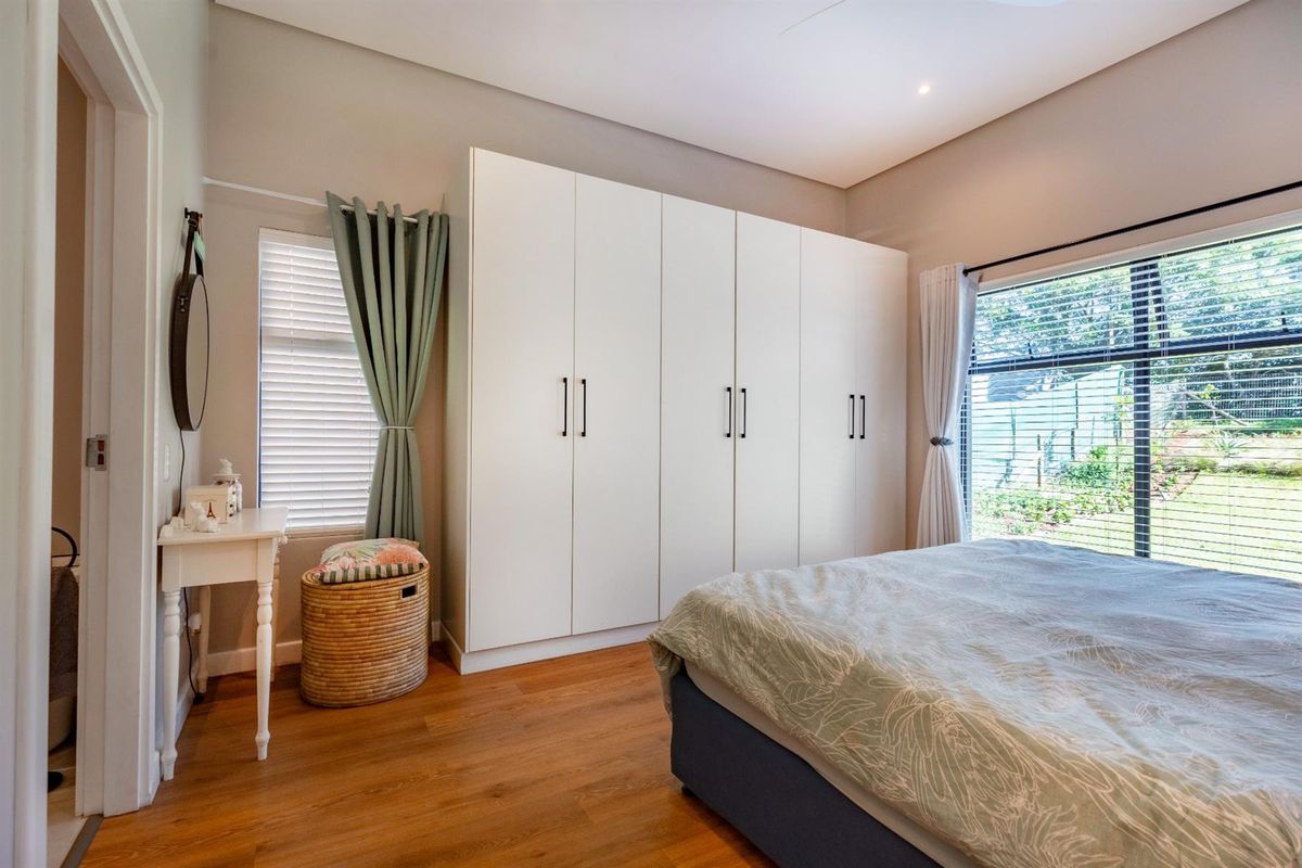 A bright bedroom featuring modern built-in floor-to-ceiling wardrobes, warm wood-look flooring, and large windows providing natural light and garden views.