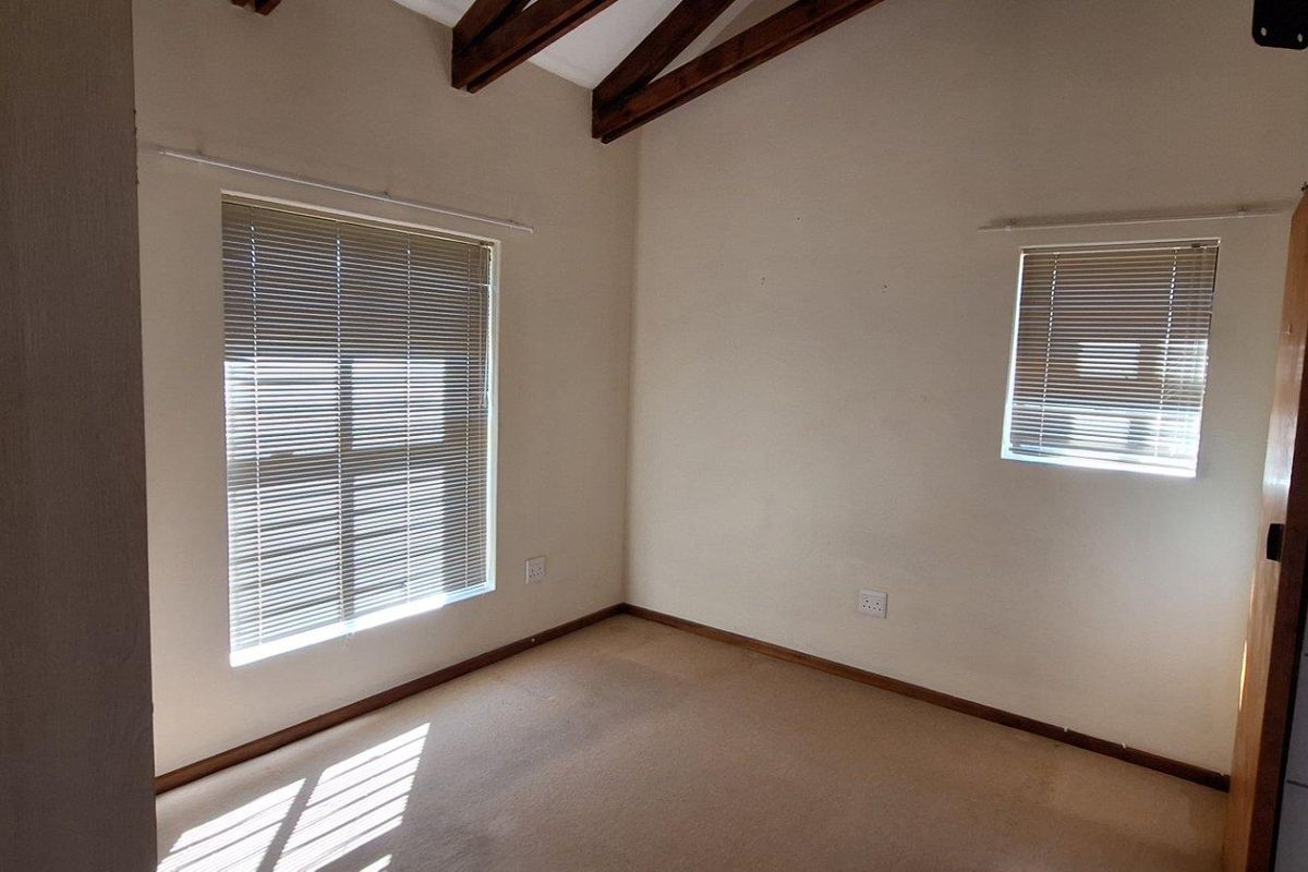 A vacant bedroom featuring exposed timber ceiling beams, carpeted flooring, and two windows fitted with venetian blinds.