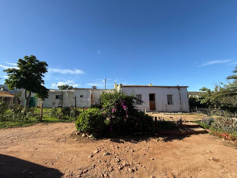 A view of the rear exterior of a single-story house featuring a weathered white facade, simple window frames, and a large garden area.