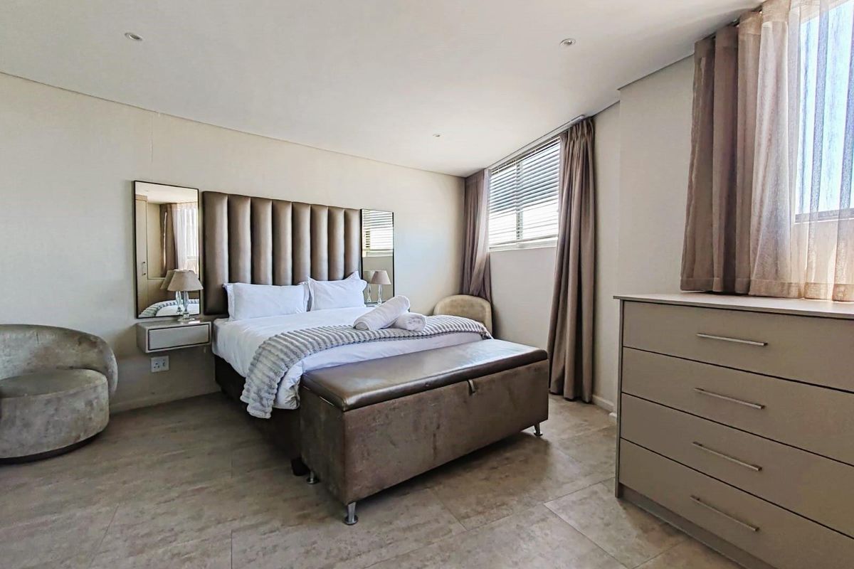 A well-maintained bedroom featuring neutral tiled flooring, recessed ceiling lighting, and a built-in headboard with integrated side mirrors and floating bedside tables.