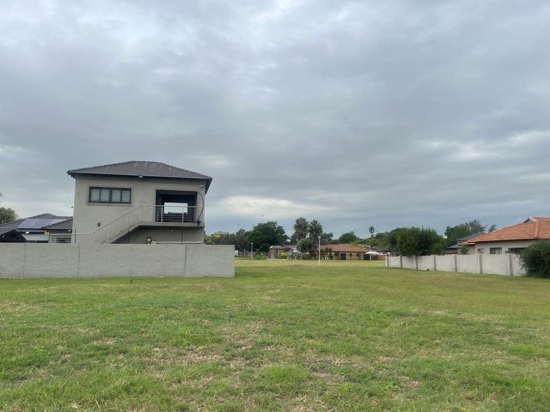 A spacious vacant residential plot featuring a well-maintained lawn, perimeter boundary walls, and an adjacent multi-level structure.