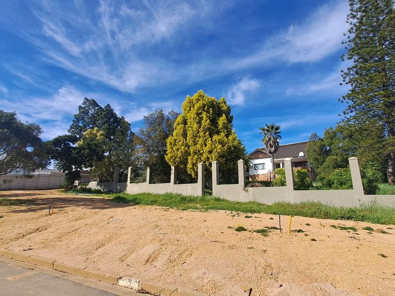 A vacant residential plot featuring a perimeter wall with concrete pillars, offering clear street access and established trees.