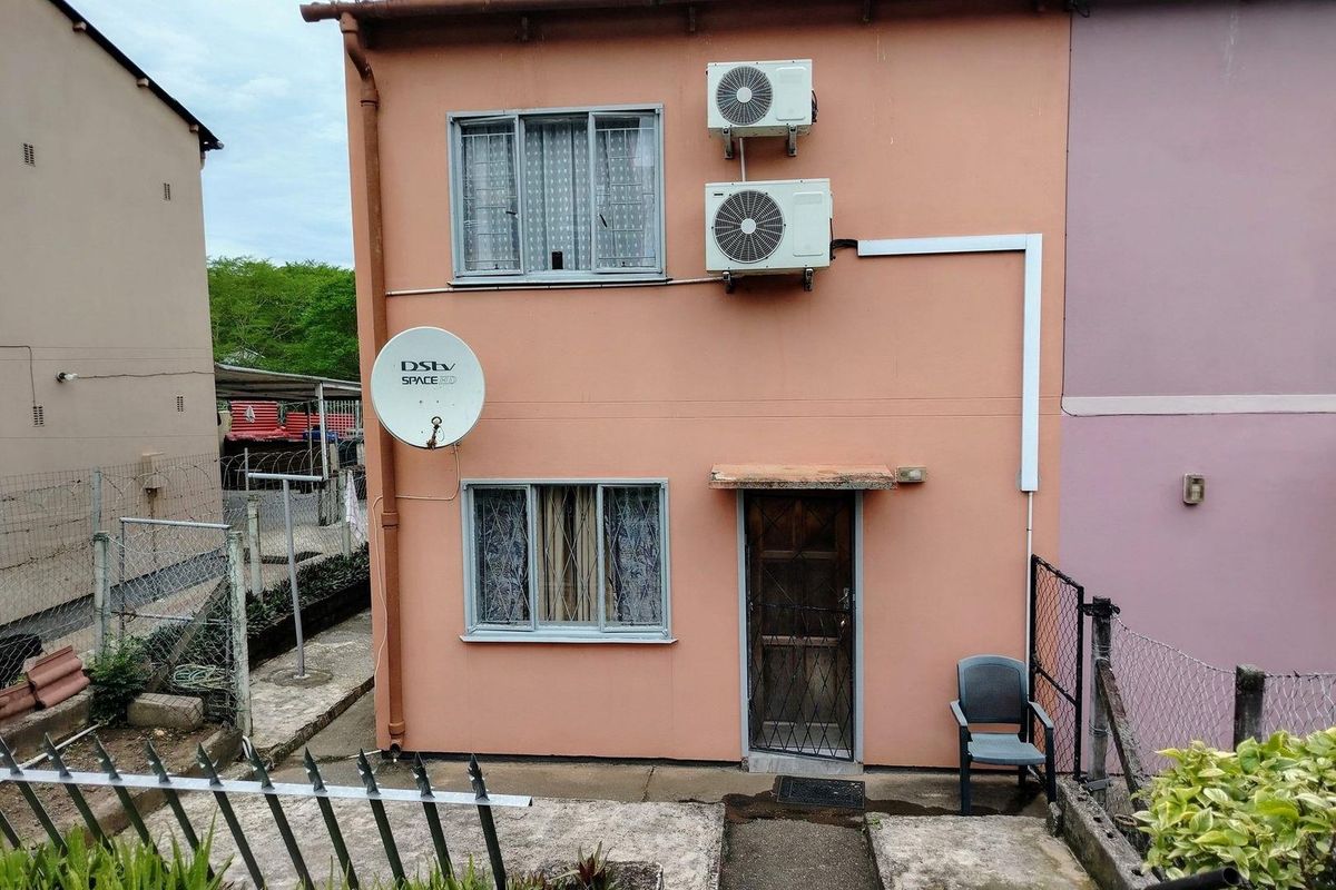 Rear exterior view of a townhouse featuring peach-toned walls, two air conditioning units, security bars on windows, and a small concrete patio area.