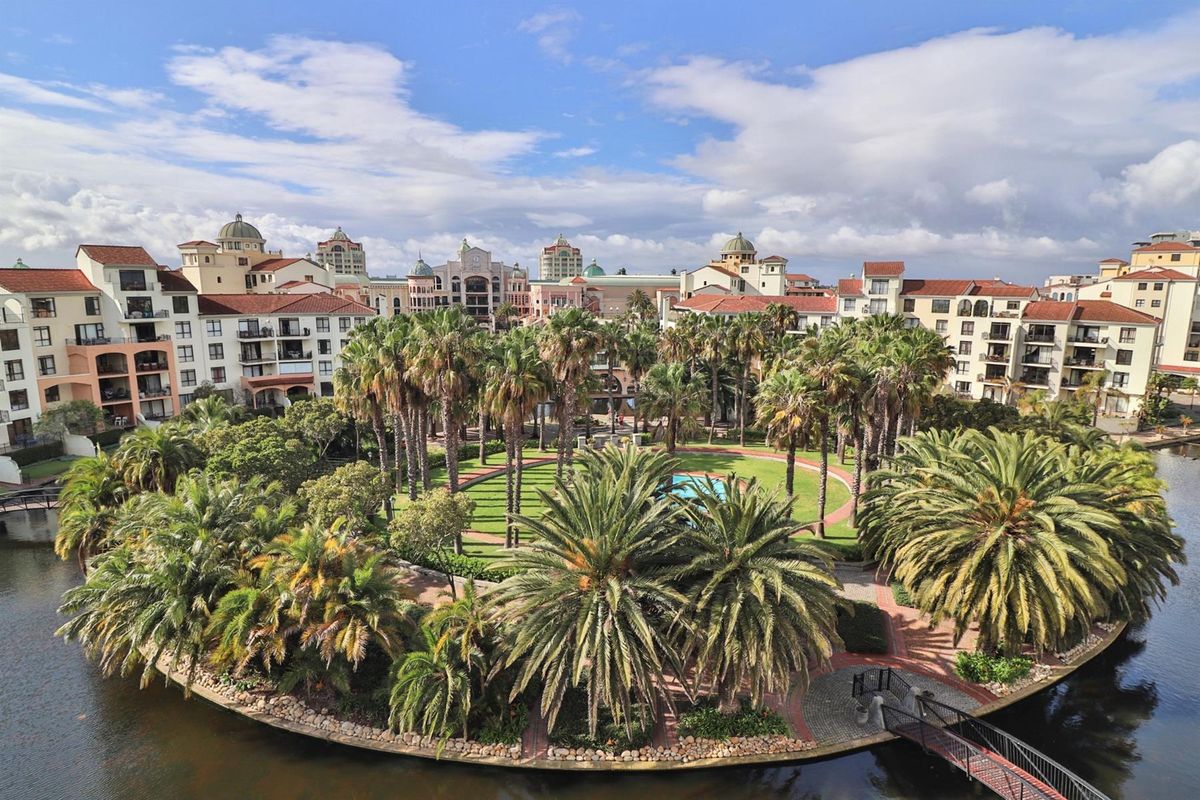 An elevated aerial view of a landscaped residential estate featuring lush palm trees, manicured lawns, a central pool area, and surrounding apartment buildings.