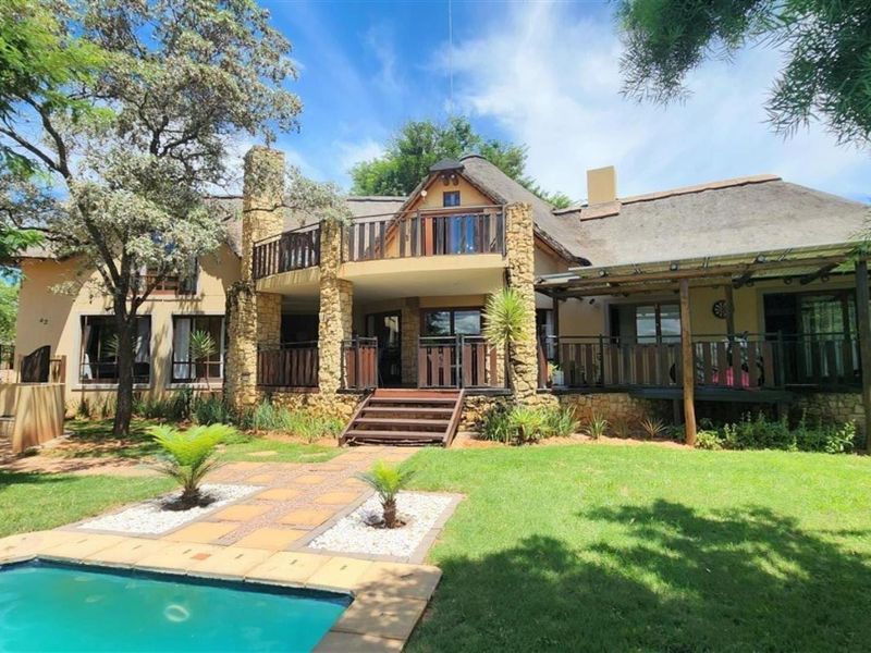 Rear view of a double-story house featuring a thatched roof, stone accents, a balcony, timber decking, and a swimming pool set in a landscaped garden.