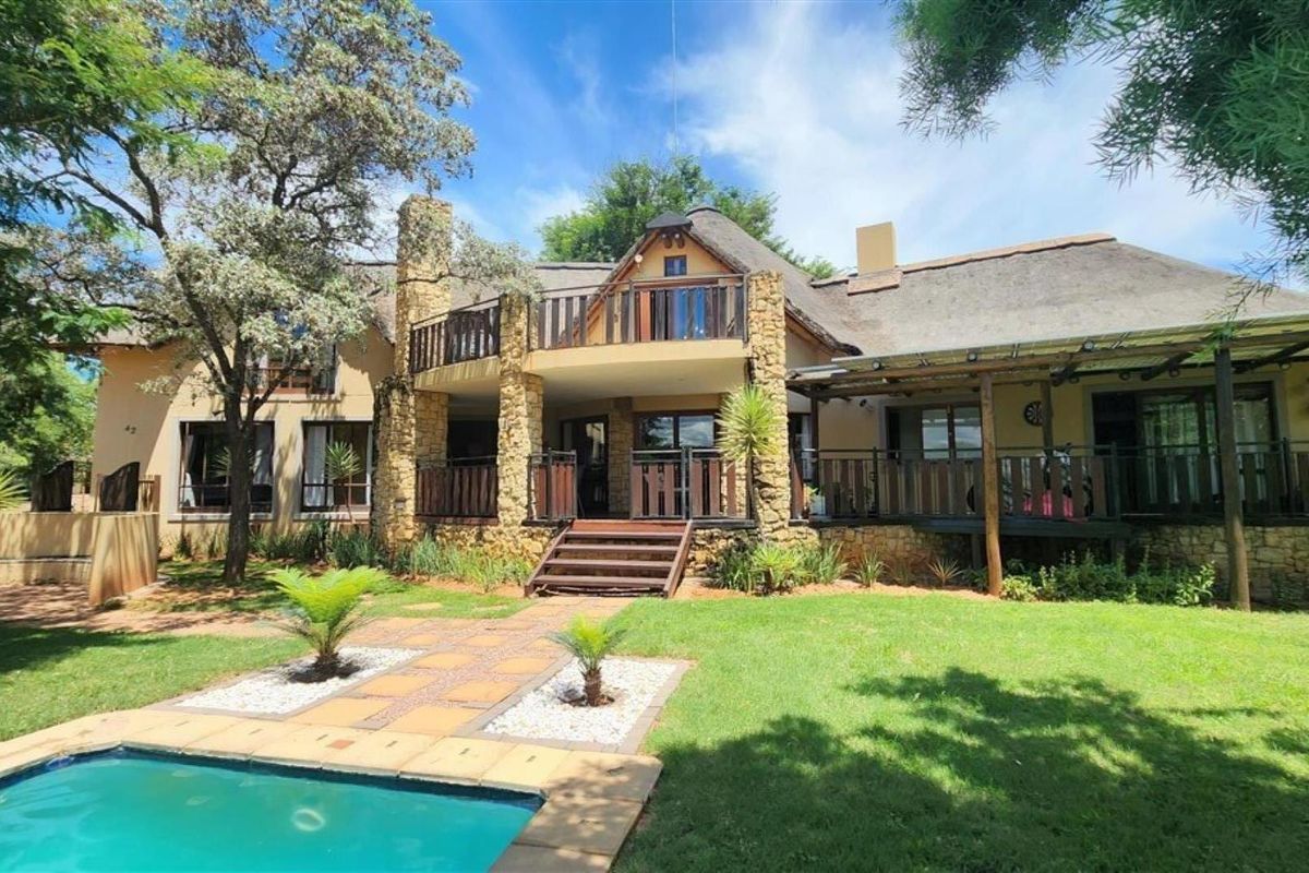 Rear view of a double-story house featuring a thatched roof, stone accents, a balcony, timber decking, and a swimming pool set in a landscaped garden.