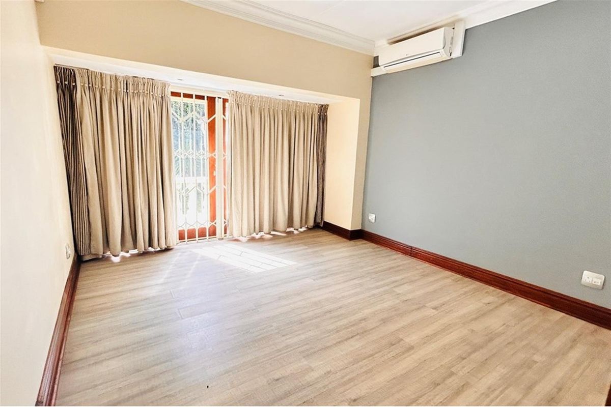 A bright, empty bedroom featuring light wood-look flooring, a wall-mounted air conditioning unit, and a sliding door with security gate access to the exterior.
