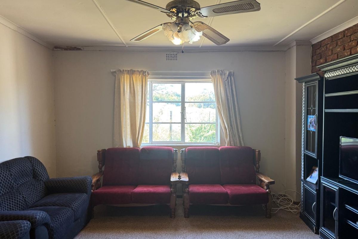 A living room featuring a ceiling fan, exposed brick accents, and a window, requiring cosmetic updates to the walls and flooring.