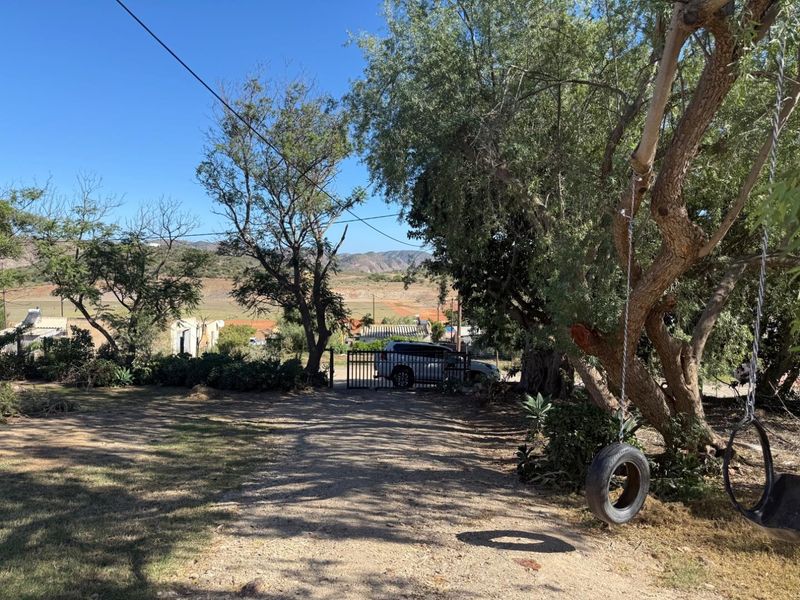 A view of the property entrance featuring a gravel driveway, metal security gate, and mature trees with a rural landscape backdrop.