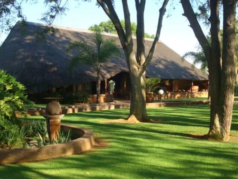 A well-maintained rear garden featuring a large thatched-roof structure, mature trees, manicured lawn, and a stone-bordered water feature.