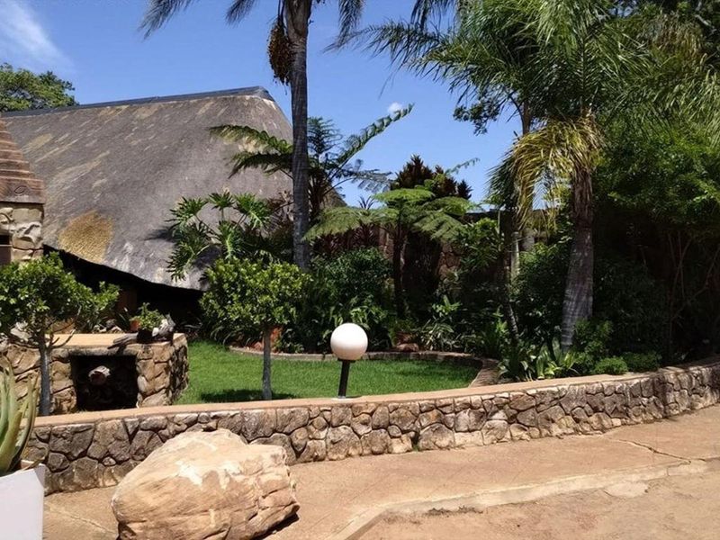 A lush garden area featuring a stone masonry wall, a built-in stone braai, and a traditional thatched roof structure set within a landscaped environment.