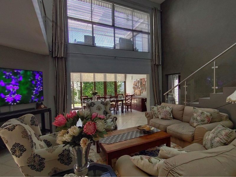 Spacious open-plan living and dining area featuring double-volume ceilings, large windows with blinds, and a modern staircase with glass balustrades.