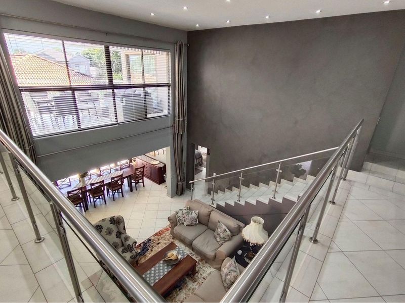 Spacious open-plan living area with double-volume ceilings, tiled flooring, and a modern staircase featuring glass and stainless steel balustrades.