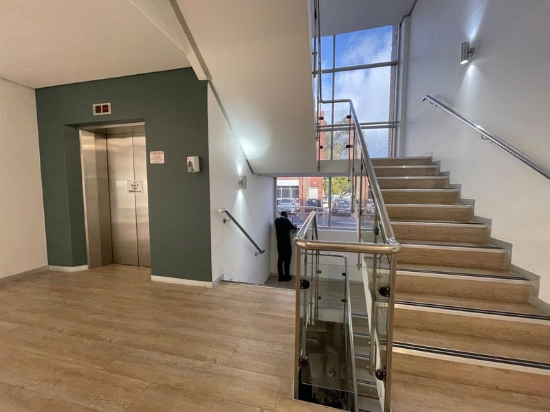 Modern office building lobby featuring a staircase with glass balustrades, wood-look tile flooring, and an elevator access point.