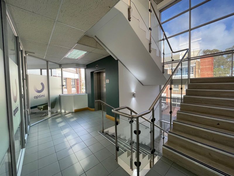 Modern office building entrance lobby featuring a glass-enclosed staircase, elevator access, and large windows providing ample natural light.