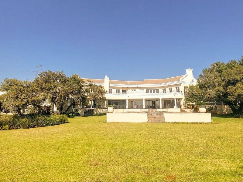 A spacious rear view of the property featuring a large manicured lawn, a stone retaining wall, and a covered patio with an upper-level balcony.