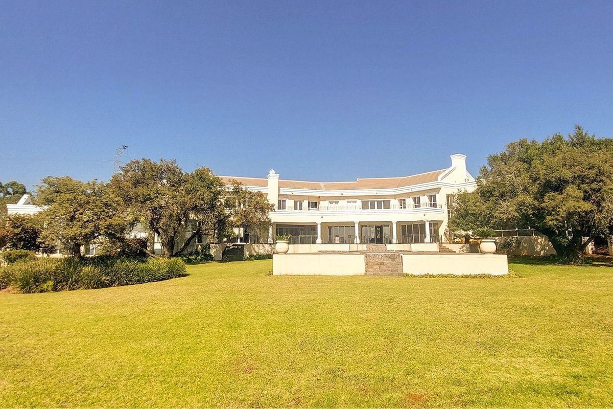 A spacious rear view of the property featuring a large manicured lawn, a stone retaining wall, and a covered patio with an upper-level balcony.