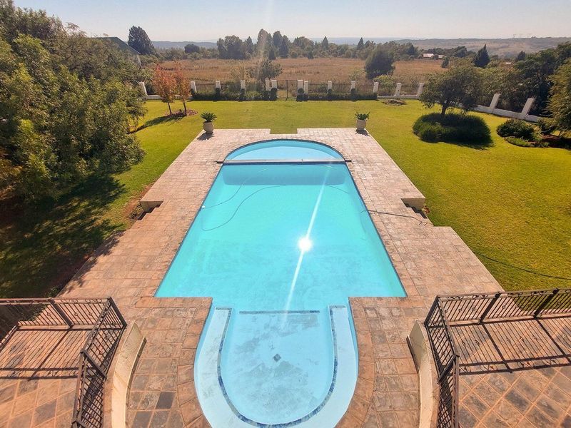 An elevated view of a well-maintained swimming pool with stone decking, surrounded by a spacious landscaped garden and perimeter fencing.