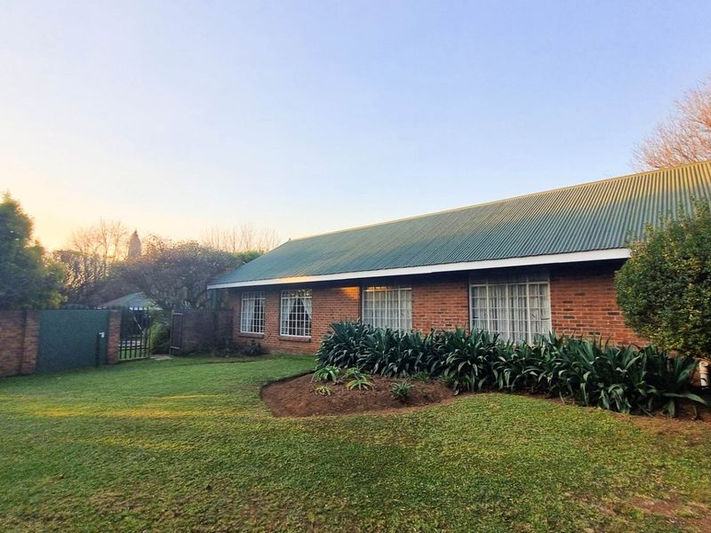 A view of the exterior rear of the brick house featuring a green corrugated metal roof, landscaped garden beds, and a lawn area.
