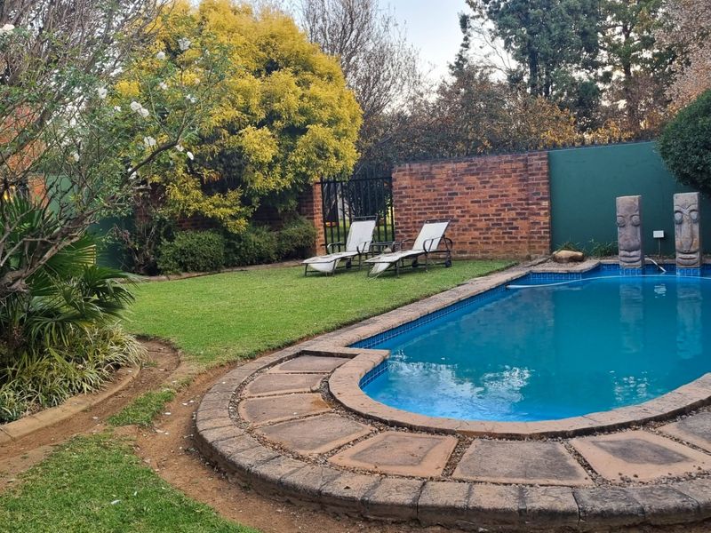 A private backyard featuring a swimming pool with stone coping, a well-maintained lawn, and established garden landscaping enclosed by a brick boundary wall.
