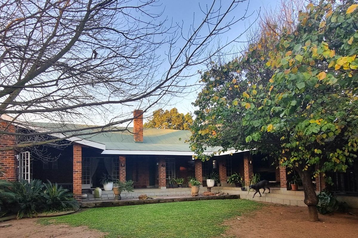 A spacious rear exterior view featuring a covered brick patio, corrugated metal roof, and established garden with mature trees.