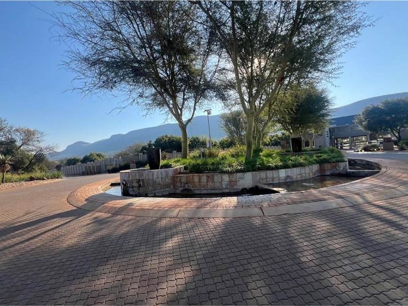 A well-maintained paved entrance roundabout featuring mature trees, stone landscaping, and a central water feature set against a scenic mountain backdrop.