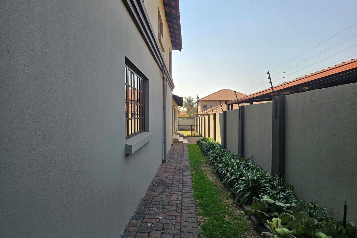 A well-maintained side exterior walkway featuring paved pathways, manicured garden borders, and secure perimeter walling with electric fencing.