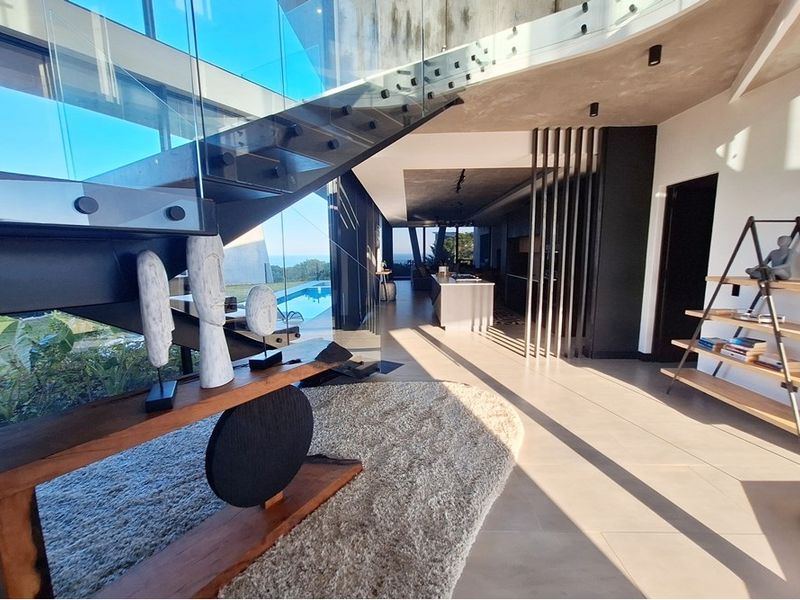 Modern open-plan living area featuring a striking floating staircase with glass balustrades, sleek tiled floors, and a seamless transition to the kitchen and pool area.