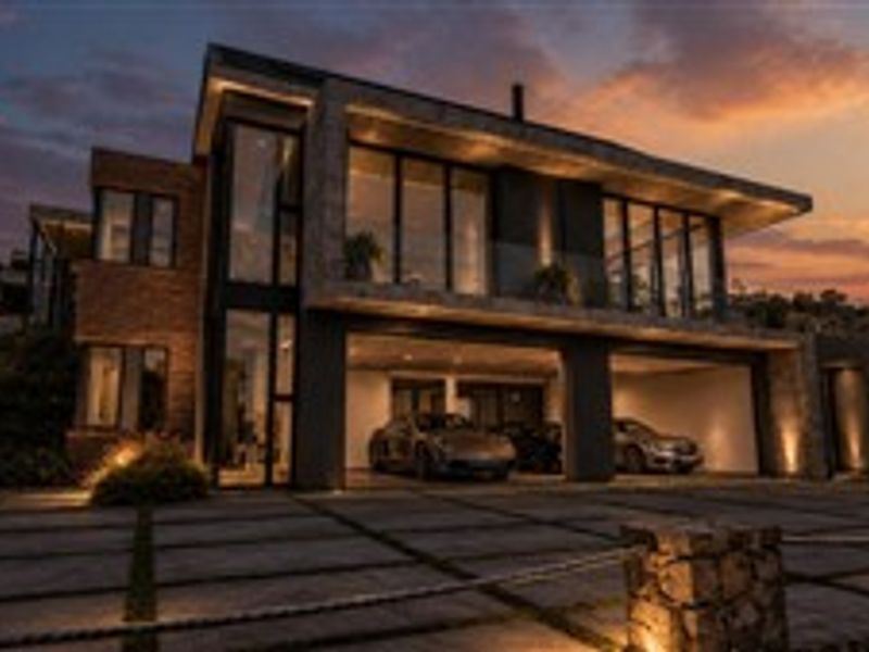 A modern luxury residence at dusk, featuring a spacious double garage, stone-paved driveway, and expansive glass facade with integrated exterior lighting.