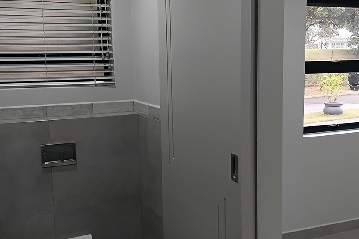A modern, neutral-toned bathroom featuring a wall-mounted flush plate, sliding door, and venetian blinds, offering a clean and contemporary aesthetic.