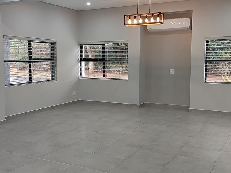 A bright, open-plan living area featuring neutral tiled flooring, modern recessed lighting, a stylish pendant fixture, and a wall-mounted air conditioning unit.