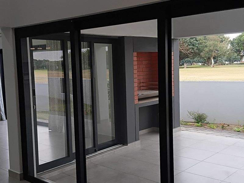 A modern covered patio featuring tiled flooring, large sliding glass doors, and a built-in brick braai area overlooking the garden.