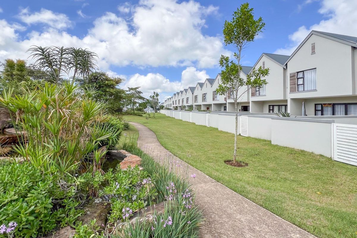 A well-maintained communal garden area featuring a paved walkway, landscaped greenery, and a water feature, adjacent to a row of modern residential apartment units.
