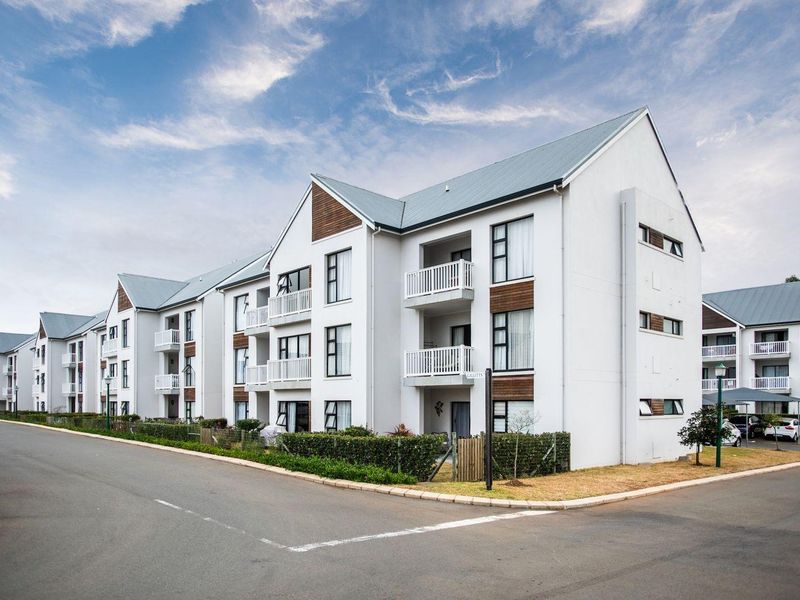 A modern multi-story apartment complex featuring pitched metal roofing, white facade finishes, timber accents, and private balconies overlooking a paved street.