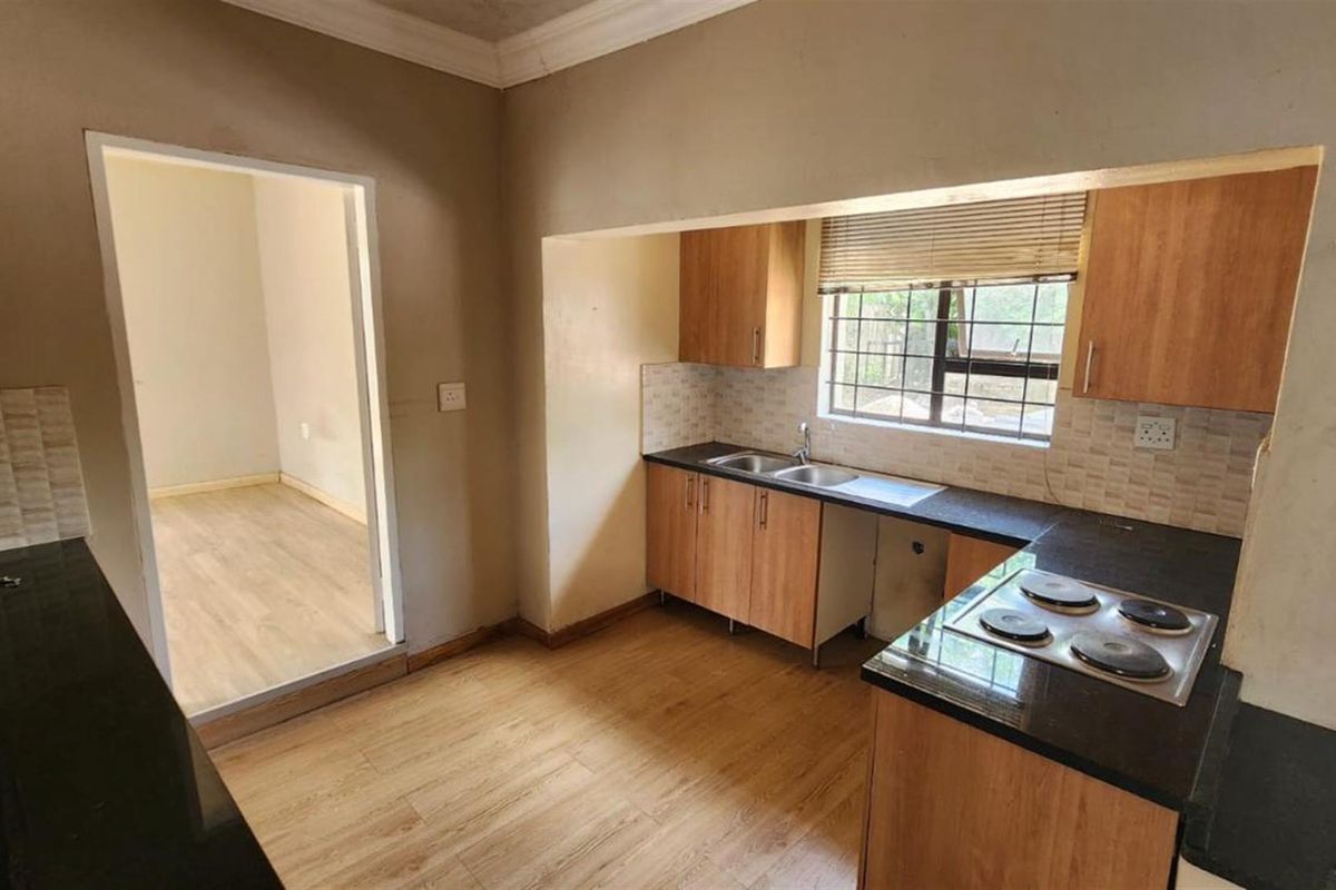 A functional kitchen featuring wood-finish cabinetry, dark countertops, an integrated electric hob, and a double sink with a view to the adjacent room.