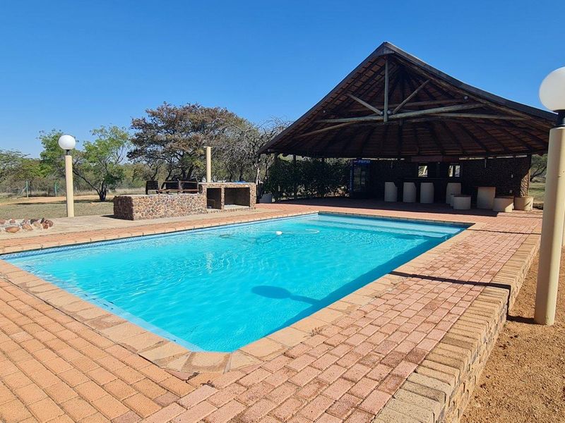 Outdoor pool area featuring a swimming pool, brick paving, and a thatched lapa with a built-in braai area, set in a rural farm landscape.