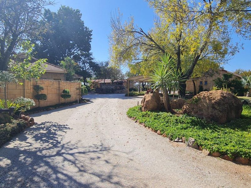 A gravel driveway leading to the property entrance, featuring landscaped garden beds, mature trees, and stone boundary walls.