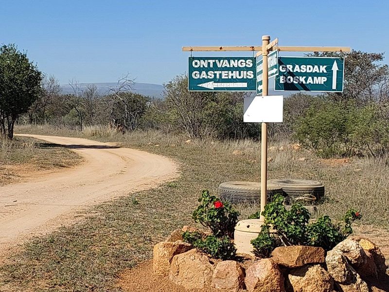 A directional signage post located on a dirt farm road, indicating the way to the guest house and bush camp.