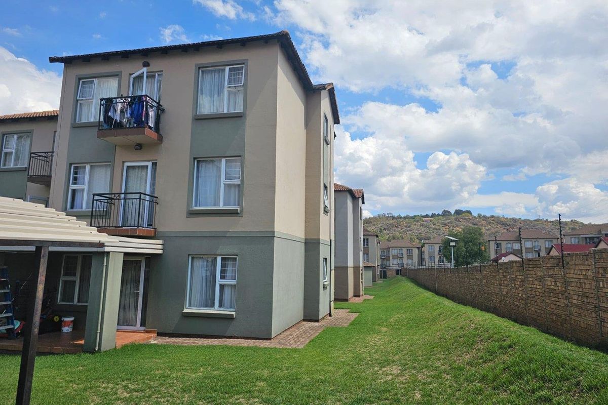 Rear view of a multi-story townhouse complex featuring balconies, a paved patio area, and a well-maintained lawn enclosed by a brick boundary wall.