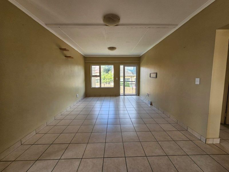 An empty, open-plan living area featuring neutral tiled flooring, ceiling lighting, and access to an outdoor space through a glass door.
