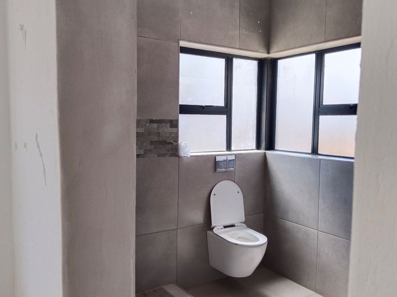A modern bathroom featuring a wall-mounted toilet with a concealed cistern, grey tiled walls, and corner windows with aluminium frames.