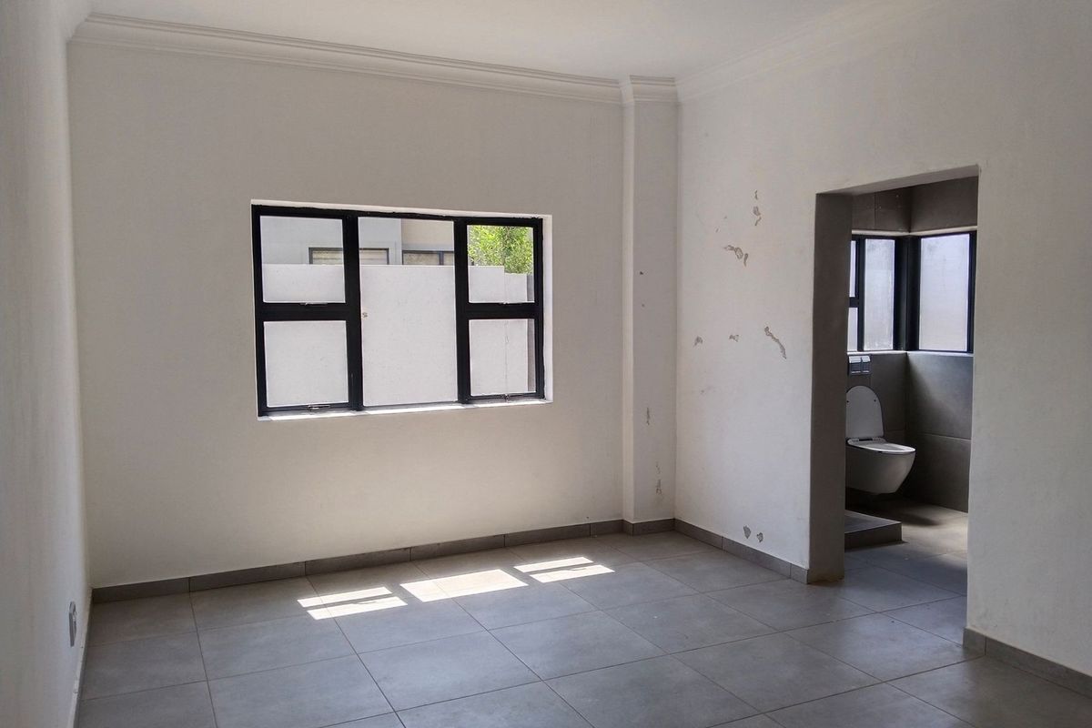 An empty bedroom featuring tiled flooring, aluminium window frames, and an attached en-suite bathroom with a wall-mounted toilet.
