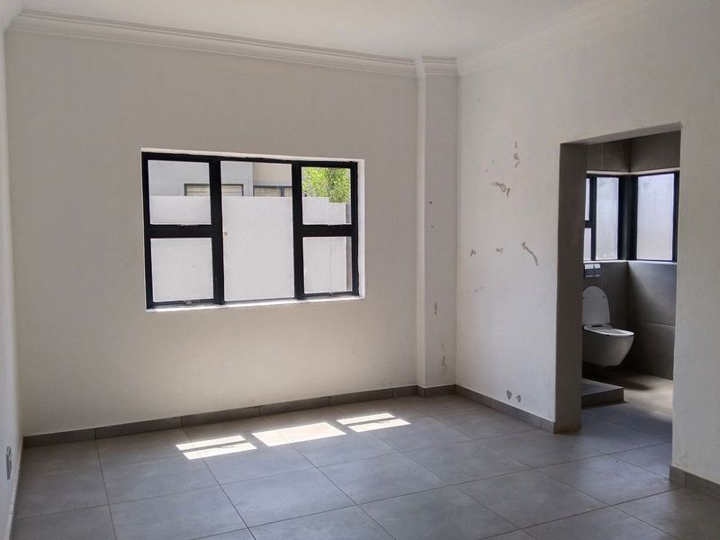 An empty bedroom featuring tiled flooring, aluminium window frames, and an attached en-suite bathroom with a wall-mounted toilet.